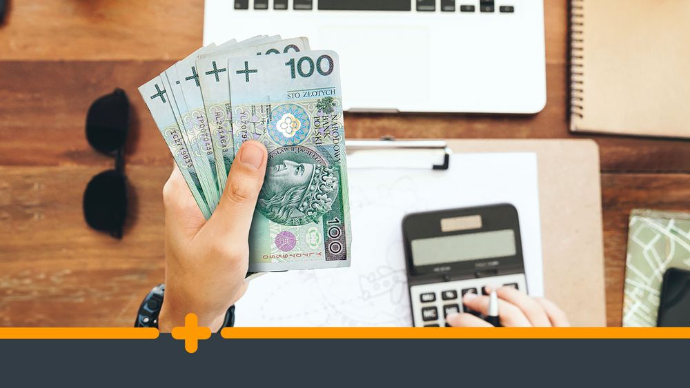 Osoba trzymająca w ręce banknoty 100 zł przy biurku z laptopem i kalkulatorem